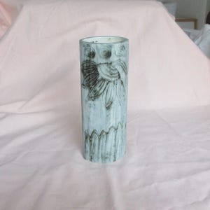 May include: A cylindrical ceramic vase featuring a light blue and white mottled glaze. The surface is decorated with dark, etched line drawings of a large flower bloom near the top, three circular motifs, and a picket fence pattern at the base.