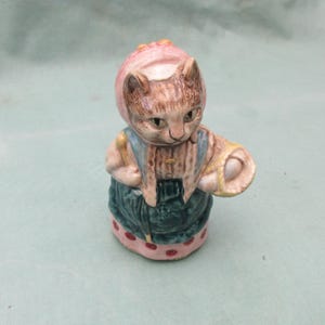 May include: A ceramic cat figurine wearing a pink bonnet, blue overalls, and a green skirt with red polka dots. The cat is light brown and holds a basket. The figurine is a decorative item.