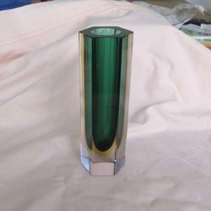 May include: A tall, hexagonal glass vase featuring a deep emerald green interior core encased in clear glass. The geometric design has six flat vertical sides and a thick, solid base, creating a layered colour effect with yellow-tinted edges.