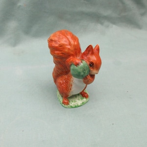 May include: A ceramic squirrel figurine with bright orange fur and a white underbelly. The squirrel clutches a green nut and stands on a patch of green grass. A delightful ornament, perfect for adding a touch of nature to any room.