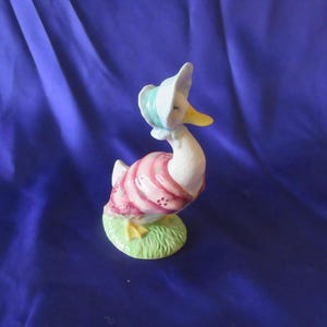 May include: A ceramic goose figurine, white with a pink and white striped dress and a blue bonnet. The goose stands on a patch of green grass, with yellow feet and beak. The figurine has a vintage, whimsical design, reminiscent of a storybook character.