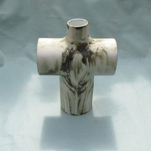 May include: A white ceramic cross-shaped vase with a brown glaze pattern. The vase has a square opening at the top.