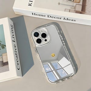 Könnte beinhalten: Transparente Handyhülle mit verspiegeltem Rücken und einem kleinen gelben Smiley. Die Hülle liegt neben zwei Büchern, eines mit dem Titel "Creative Living" und das andere "Home Decor Ideas."