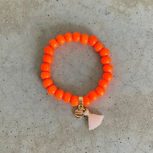 Könnte beinhalten: Ein leuchtend orangefarbenes Perlenarmband mit einem goldenen Anhänger, auf dem "Kiss My Wish" steht, und einem rosa Quaste.