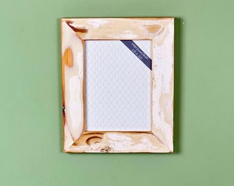 Peut inclure: Un cadre photo rectangulaire en bois avec une finition vieillie blanc cassé. Le cadre a un grain de bois clair et un support texturé blanc. Une étiquette bleu foncé dans le coin indique "Marzunikat 15 x 20". Le cadre est accroché à un mur vert.