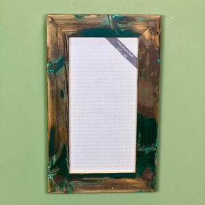 Peut inclure: Un cadre photo rectangulaire en bois patiné avec une finition vieillie dans des tons marron et vert foncé. Le cadre contient un insert blanc à motif répétitif et une étiquette diagonale portant l'inscription Harzunikat 15 x 30.