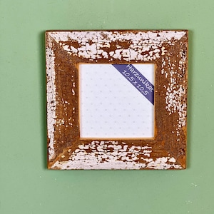 Può includere: Cornice quadrata in legno con finitura bianca anticata su base marrone. La cornice ha una visibile venatura del legno e una piccola etichetta blu con testo. Dimensioni: 10,5 x 10,5 cm.