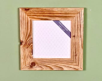 Peut inclure: Cadre photo carré en bois clair avec des nœuds apparents. Une étiquette violette dans le coin indique "Harzunikat 15 x 15". Le cadre est posé sur un fond vert.