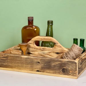 Könnte beinhalten: Ein rechteckiger Holzkasten mit einem mittigen Griff und einer dunklen, geflammten Holzmaserung. Im Kasten befinden sich eine braune Glasflasche, eine grüne Glasflasche, zwei kleine grüne Glasflaschen, ein Holzwerkzeug und ein Knäuel braunes Garn.