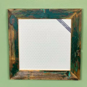Può includere: Cornice quadrata in legno con finitura rustica e anticata. La cornice è dipinta con sfumature di verde e marrone, con venature del legno visibili. Interno bianco e un'etichetta con la scritta "Harzunikat 25 X 25".