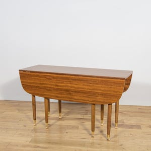 Könnte beinhalten: Ein Mid-Century-Modern-Klapptisch mit warmem Holzmaserungs-Finish. Der Tisch hat vier schlanke, sich verjüngende Beine mit messingfarbenen Spitzen. Der Tisch ist etwa 76 cm lang, wenn die Blätter unten sind.