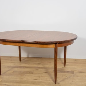 Peut inclure: Une table à manger ovale dotée d'un plateau en bois brun foncé au grain apparent avec une couture centrale visible, reposant sur quatre pieds en bois clair effilés. La table présente un bord arrondi lisse et une structure simple.