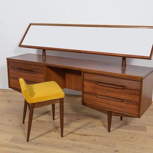 Op de afbeelding: Mid-century modern houten kaptafel met een rechthoekige spiegel en een gele kruk. De kaptafel heeft lades en een open middenruimte. Het hout is bruin en de kruk heeft een gebogen zitting. De tafel is ongeveer 150 cm breed.