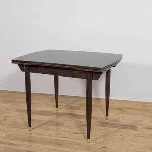 Peut inclure: Table de style moderne du milieu du siècle, de couleur marron foncé, avec un plateau carré et quatre pieds effilés. Les pieds ont des embouts dorés. La table mesure environ 76 cm de long, 76 cm de large et 74 cm de haut.