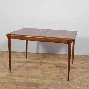 May include: A rectangular wooden dining table with a smooth, medium-brown finish. The table features a flat top surface with a visible seam in the centre, indicating an extendable design, supported by four slender, tapered legs at each corner.