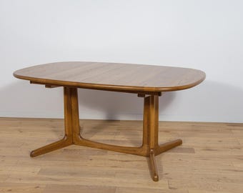 Può includere: Tavolo da pranzo in legno marrone chiaro con piano ovale e due set di gambe. Il tavolo è in legno e ha una finitura naturale. Le gambe hanno un design unico, con una base curva e un supporto verticale.