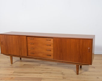 May include: A long, rectangular, mid-century modern teak sideboard with two sliding doors and a set of drawers in the center. The sideboard has a light wood finish and sits on four tapered legs.