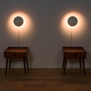 Puede incluir: Dos mesitas de noche a juego de estilo moderno de mediados de siglo, con cajones y patas cónicas. Cada mesa se combina con una lámpara de pared redonda gris. Las lámparas proyectan un brillo cálido en la pared. Las mesas son de madera.