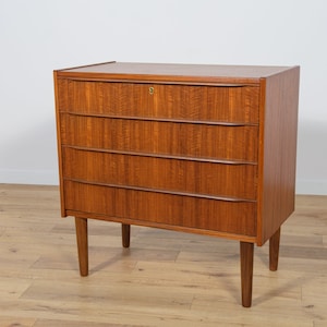 Può includere: Un cassettone a quattro cassetti in legno marrone chiaro con un buco della serratura sul cassetto superiore. Il cassettone ha un design semplice con bordi arrotondati e poggia su quattro gambe corte.