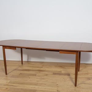 Może przedstawiać: Owalny stół jadalniany w stylu mid-century modern z ciepłym brązowym wykończeniem. Stół ma gładką powierzchnię i cztery zwężane nogi. Stół ma około 183 cm długości i 102 cm szerokości.