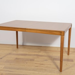 May include: A rectangular wooden dining table with a light brown tabletop and four slender, tapered legs. The table is set against a white wall and sits on a light brown wooden floor. The table is approximately 60 inches long.