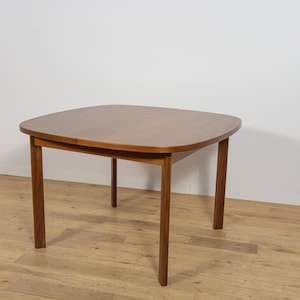 Peut inclure: Une table à manger carrée en bois avec des coins arrondis et une finition brun moyen lisse. La table présente quatre pieds droits et effilés ainsi qu'une couture visible au centre du plateau, indiquant une conception extensible.
