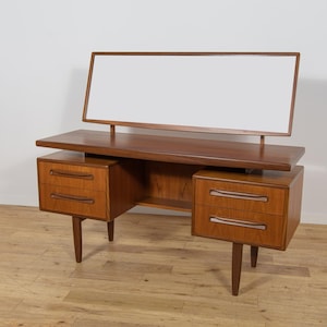 Op de afbeelding: Een kaptafel van teakhout met een rechthoekige spiegel aan de achterzijde. Het bureau heeft een vlak bovenblad en twee sets van twee lades aan weerszijden, elk voorzien van horizontale houten handgrepen en rustend op vier taps toelopende poten.