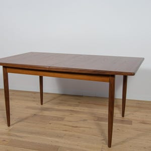 Può includere: Un tavolo da pranzo in legno in stile moderno di metà secolo con un piano rettangolare e quattro gambe. Il tavolo è in legno scuro e ha una finitura naturale.