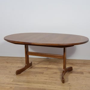 Könnte beinhalten: Ein ovaler Esstisch im Mid-Century-Modern-Stil. Der Tisch ist aus Holz mit warmbraunem Finish gefertigt. Der Tisch hat ein einzigartiges Basisdesign mit gebogenen Beinen. Der Tisch ist etwa 152 cm lang und 102 cm breit.