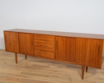 May include: A long, rectangular, mid-century modern teak sideboard with four drawers and two sets of sliding doors. The sideboard has a warm, honey-brown color and is sitting on a light wood floor.