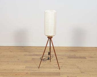 Könnte beinhalten: Eine weiße Stehlampe mit einem zylindrischen Schirm und einem dreibeinigen Sockel aus dunklem Holz.