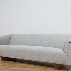 Peut inclure: Un canapé gris clair avec un dossier et des accoudoirs incurvés. Le canapé a un profil bas et repose sur des pieds en bois. Le revêtement semble être un tissu doux et texturé. Le canapé est de couleur neutre et s'intégrerait dans de nombreux styles de décoration.