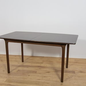 Peut inclure: Une table à manger rectangulaire avec une finition en bois brun foncé, dotée d'une surface supérieure lisse et plate et de quatre pieds fins légèrement effilés qui s'étendent depuis les coins. La table présente une couture visible au centre.