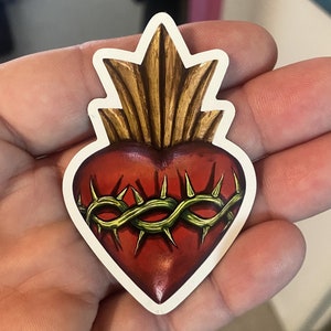 May include: A red heart with a gold crown and a green thorn crown around it. The heart is a religious symbol.