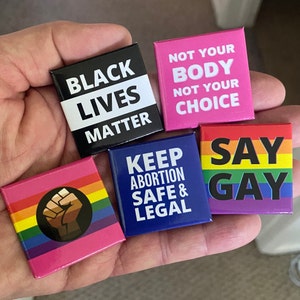 May include: Five square buttons with different messages and colours. The buttons are black, pink, blue, and rainbow. The messages on the buttons are "Black Lives Matter", "Not Your Body Not Your Choice", "Keep Abortion Safe & Legal", "Say Gay", and a black fist on a rainbow background.