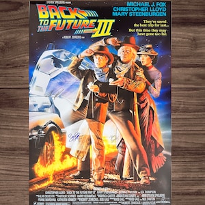 May include: A movie poster for Back to the Future Part III, featuring Michael J. Fox, Christopher Lloyd, and Mary Steenburgen. The poster is set in the Wild West, with the DeLorean time machine in the background. The poster is signed by the three actors.