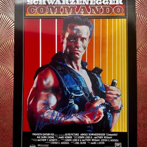 以下が含まれることがあります： アーノルド・シュワルツェネッガー主演の映画「コマンドー」の額入りポスター。ポスターは、上部にタイトルが付いた赤と黄色のストライプの背景です。シュワルツェネッガーは青いベストを着て、武器を持ち、彼のサインが見えます。