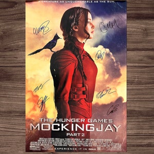 May include: A movie poster for The Hunger Games: Mockingjay Part 2. The poster features a woman in a red jumpsuit with a mockingjay bird on her shoulder. The poster is signed by the cast members. The text on the poster reads "A creature as unquenchable as the sun." and "The Hunger Games: Mockingjay Part 2".