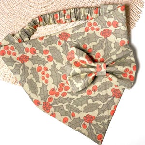 May include: A beige and green bandana with a red holly berry pattern and a matching bow tie. The bandana has a ruffled edge.