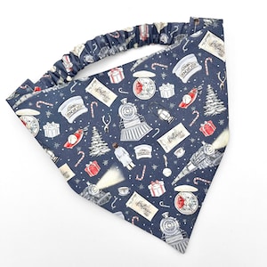 May include: A navy blue headband with an elastic band, featuring a repeating pattern of trains, Christmas trees, gifts, candy canes, and the word "Believe". The design includes conductor hats and pocket watches.