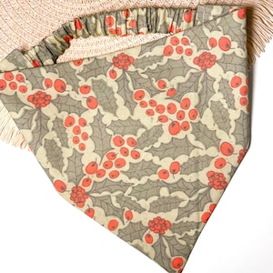 May include: A beige bandana with a red and green holly berry pattern. The bandana has a ruffled edge.