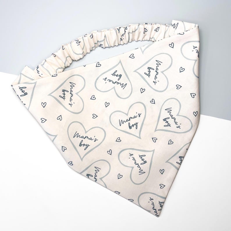 K&ouml;nnte beinhalten: Ein wei&szlig;es Bandana mit einem grauen Herzmuster und dem Text "Mama's boy", der sich wiederholt. Das Bandana hat ein Gummiband.
