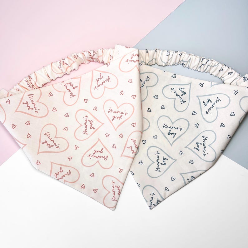 K&ouml;nnte beinhalten: Zwei wei&szlig;e Stoffbandanas mit elastischen R&auml;ndern. Ein Bandana hat ein rosafarbenes Herzmuster mit dem Text "Mama's girl" und das andere hat ein graues Herzmuster mit dem Text "Mama's boy".