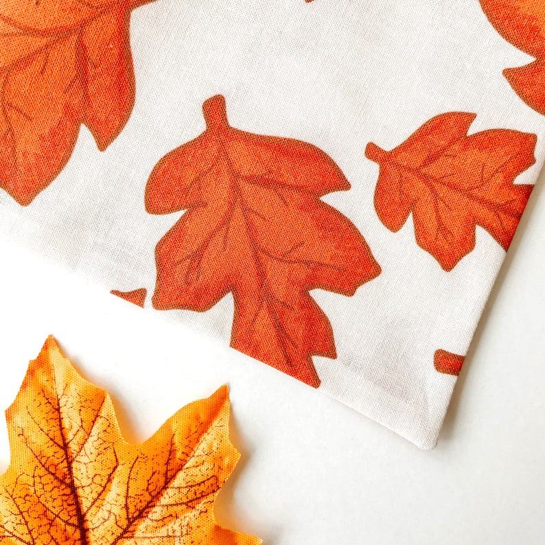 May include: A white fabric with a pattern of orange and brown leaves. The fabric is folded in half and the corner is turned up. There is a single orange leaf in the bottom right corner of the image.