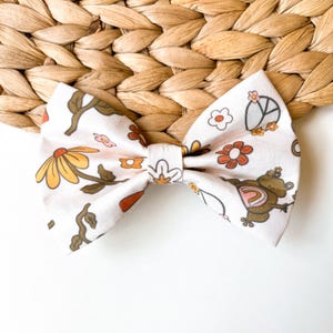 May include: A white fabric bow with a floral and frog print. The print features orange, yellow, brown, and green colors.