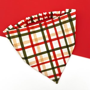 May include: A plaid bandana in red, green, and tan, with a white background. The bandana has a red and white elastic band. The pattern is a grid of intersecting lines. The bandana is triangular in shape.