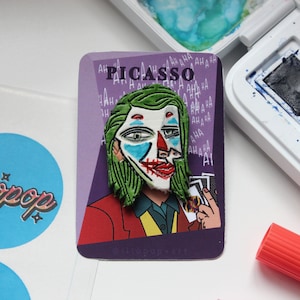 May include: A purple card with the word "PICASSO" printed in white letters. The card features a green and white cartoon image of the Joker from the movie "Joker" with the text "AHA HA HA" repeated around the image.
