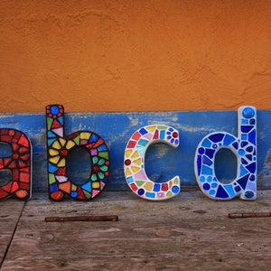 Könnte beinhalten: Vier mosaikartige Kleinbuchstaben: a, b, c und d. Die Buchstaben bestehen aus bunten Glasstücken und sind auf einer Holzoberfläche vor einem orangefarbenen und blauen Hintergrund angeordnet. Die Buchstaben sind etwa 10 cm hoch.