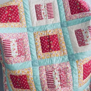 Könnte beinhalten: Eine Patchwork-Decke mit hellblauem Hintergrund und Quadraten in verschiedenen floralen und geometrischen Mustern in Rot, Rosa und Gelb.