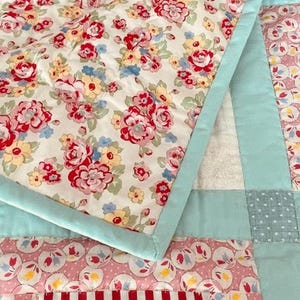 K&ouml;nnte beinhalten: Eine Patchwork-Decke mit einem Blumendruck auf der obersten Schicht. Die Decke besteht aus verschiedenen Stoffen in den Farben Rosa, Blau und Wei&szlig;. Die Decke ist mit wei&szlig;em Garn gesteppt.